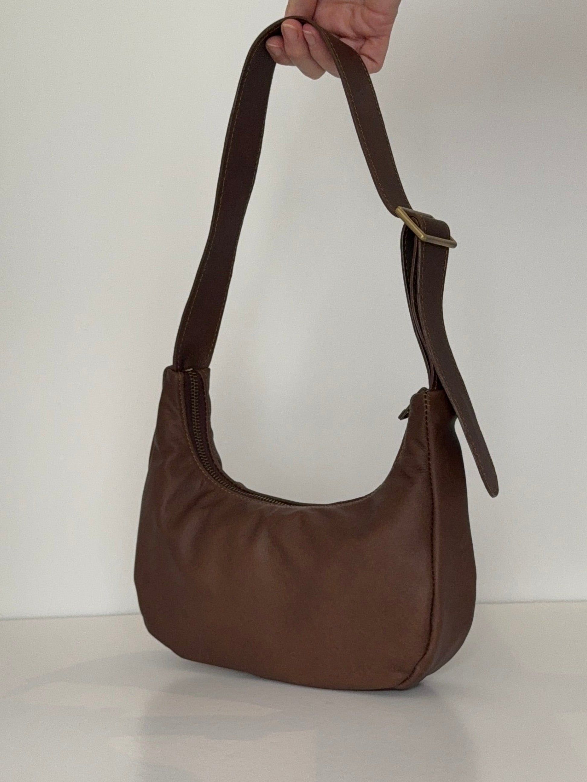 Hand holding brown baguette shaped leather bag with antique brass zip and slide