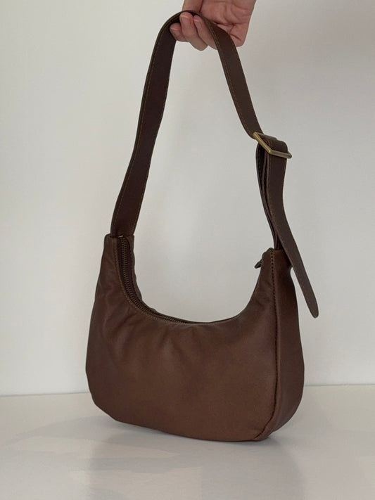 Hand holding brown baguette shaped leather bag with antique brass zip and slide